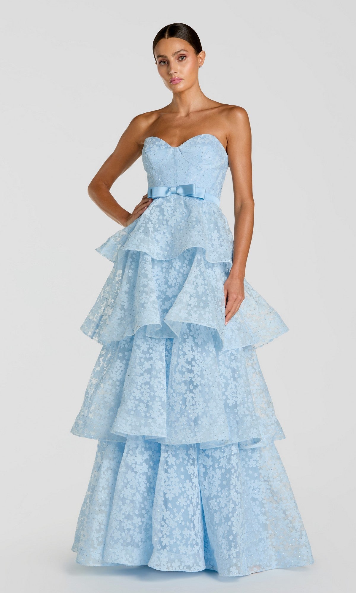 A woman wearing the Mac Duggal 56298 long formal dress in powder blue—featuring a strapless, organza layered design with a bow at the waist—stands against a plain light gray background.